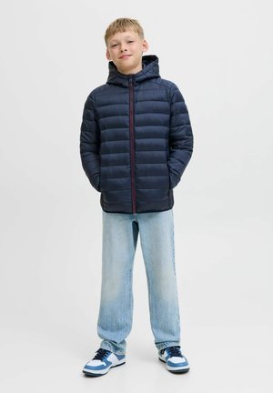 Marineblauwe gewatteerde jas met capuchon en rode rits, gecombineerd met lichtblauwe losse jeans en witte sneakers met blauwe accenten. Effen achtergrond.