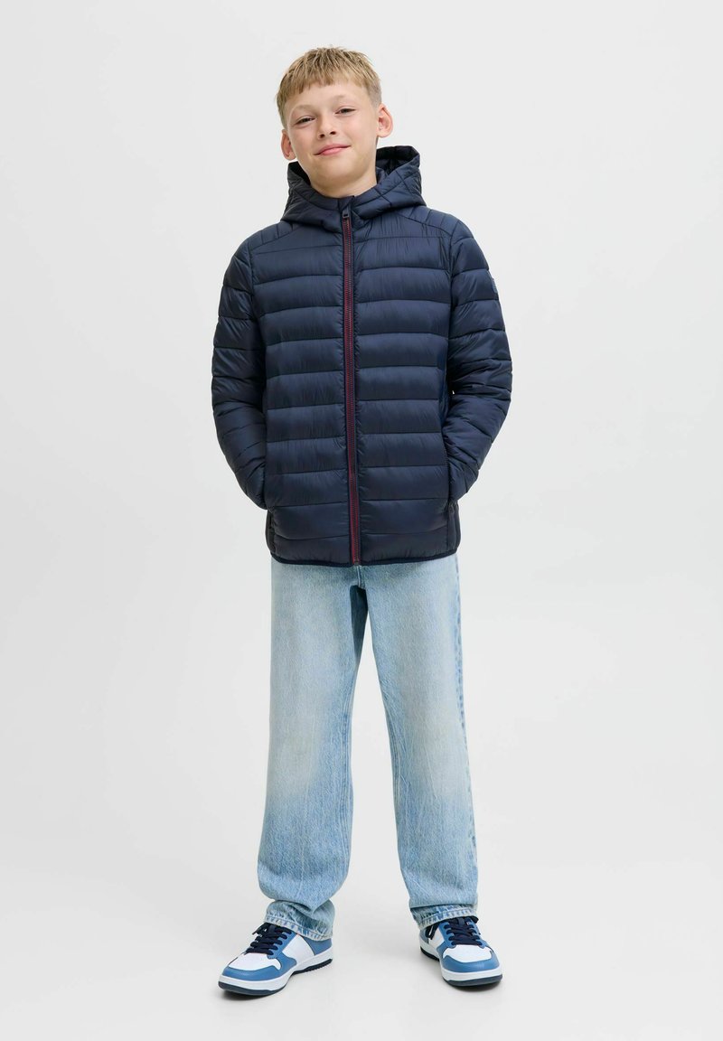 Marinblå vadderad jacka med huva och röd dragkedja, kombinerad med ljusblå löst sittande jeans och vita sneakers med blå detaljer. Enfärgad bakgrund.