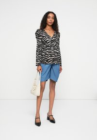 Schwarz-beiges Zebra-Muster Langarmtop, Jeansshorts, cremefarbene Handtasche und schwarze High Heels. Das Model steht vor einem weißen Hintergrund.