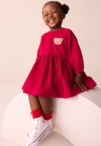 Vestito rosso con maniche lunghe, vita arricciata e applicazione di orso. Abbinato a sneakers bianche e calzini rossi, su una piattaforma bianca e arrotondata.