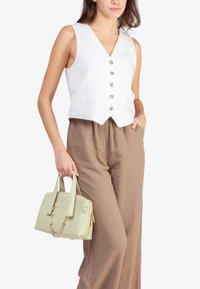 Gilet bianco in lino senza maniche con cinque bottoni, abbinato a pantaloni beige a gamba larga. Il modello tiene una borsa verde chiaro con dettagli in catena dorata.