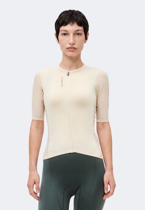 Γυναίκα με κοντά σκούρα μαλλιά φοράει μπεζ κοντομάνικη φανέλα ποδηλασίας και σκούρα πράσινα επενδύμενα ποδηλατικά σορτς, στέκεται μπροστά σε μονόχρωμο φόντο.