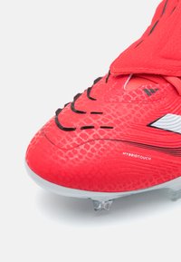 Scarpa da calcio rossa con tomaia sintetica testurizzata, caratterizzata da cuciture e dettagli neri. Tacchetti trasparenti e stampati sulla suola per una migliore trazione.