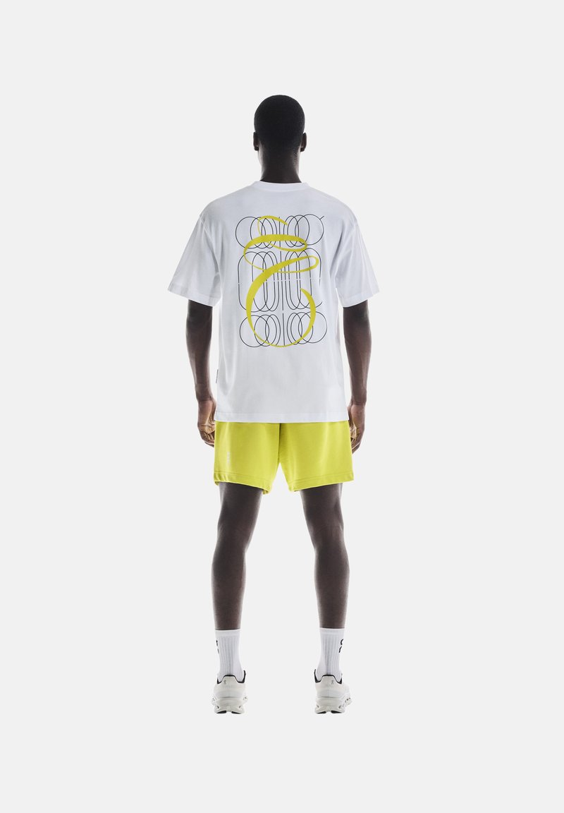 Weißes T-Shirt mit kurzen Ärmeln und einem bunten Grafikerdesign auf der Rückseite, kombiniert mit knallgelben Shorts und weißen Socken mit schwarzen Akzenten.