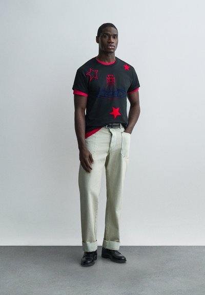 Νέος άνδρας που στέκεται σε τοίχο γκριχρώματος, φοράει μαύρο μπλουζάκι με κόκκινα αστέρια, μπεζ παντελόνι, μαύρη ζώνη και μαύρα παπούτσια.