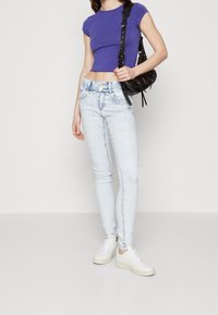 Top corto de manga corta morado, jeans skinny de lavado claro con un ligero desvanecimiento, combinados con zapatillas blancas y un pequeño bolso negro de hombro.