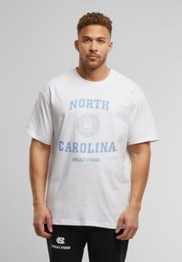 Hvid bomulds t-shirt med blå tekst "NORTH CAROLINA" og "UPSCALE STUDIOS" samt et rundt logo design. Korte ærmer, afslappet pasform.