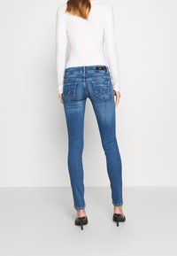 Jeans skinny blu con un lavaggio sbiadito, due tasche posteriori e dettagli di cucitura sottili, abbinati a un top bianco a maniche lunghe e tacchi neri.
