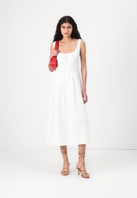 Abito bianco senza maniche con ricami floreali e gonna svasata. La modella tiene una borsa rossa. Indossato con sandali con tacco beige e cinturini.