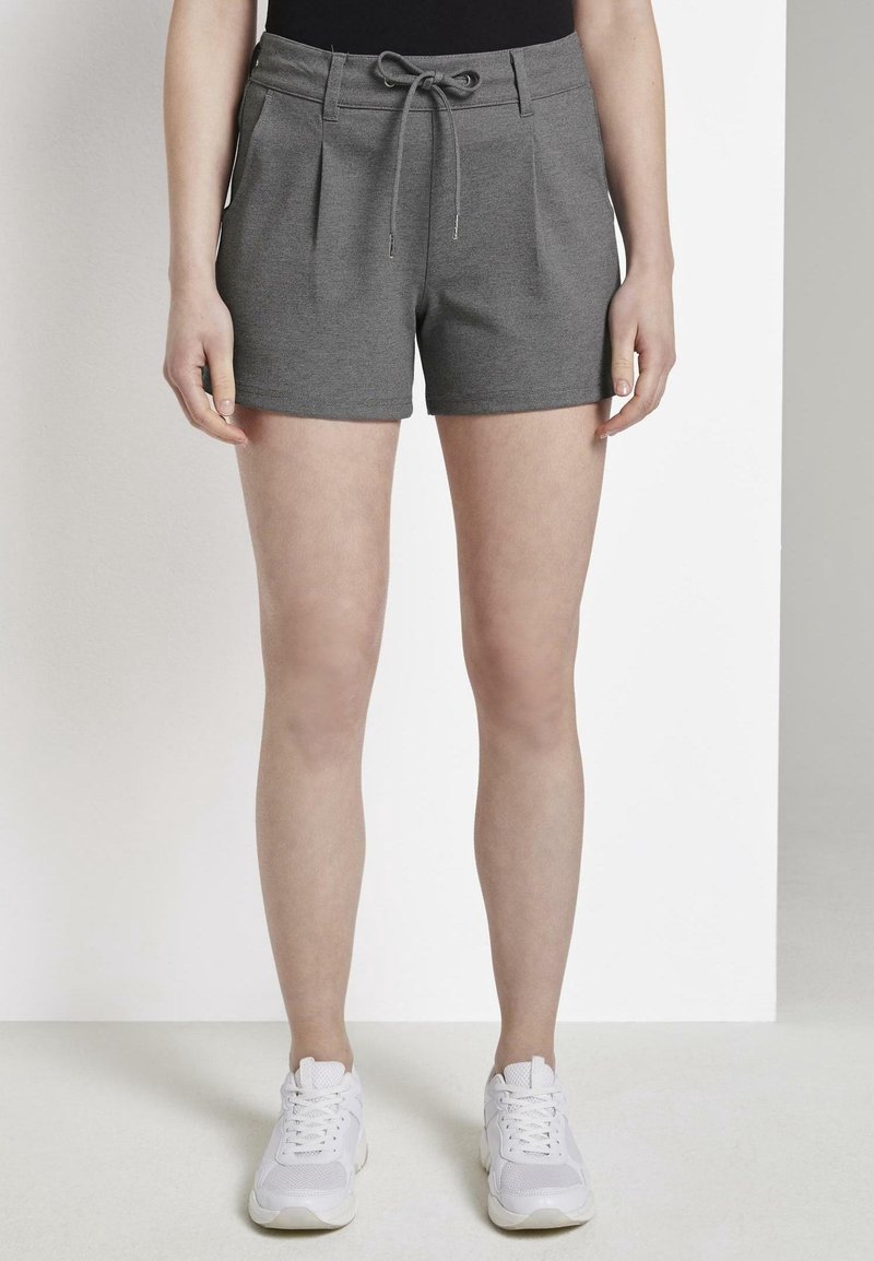 Femme portant un short gris à cordon de serrage et des baskets blanches, se tenant devant un fond blanc et gris clair uni.