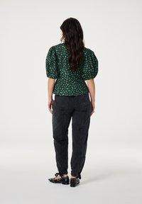 Groene top met een patroon en pofmouwen, met een getailleerde taille, gecombineerd met zwarte baggy broek. Zwarte schoenen hebben enkelbanden en metalen accenten.
