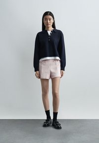 Kvinne som står mot en ensfarget vegg iført en marineblå knappet genser, hvit undertrøye, rosa teksturerte shorts, svarte sokker og svarte loafers.