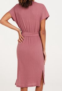 Robe midi rose framboise en tissu texturé, avec manches courtes, taille ceinturée, et fentes latérales pour faciliter les mouvements.