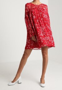 Robe florale rouge à manches longues, en tissu transparent, avec une coupe décontractée. Associée à des chaussures à talons carrés blanches, mettant en valeur le motif vibrant de la robe.