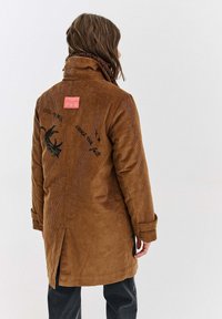 Brązowy płaszcz z kordurowej tkaniny z wysokim kołnierzem, haftowanym wzorem kota oraz napisem "ZMIENIAJ SWOJE PRZEZNACZENIE". Posiada metkę z tyłu i boczne rozcięcia.