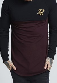 Man som bär en svart och mörkröd långärmad skjorta med guldbroderad logotyp på bröstet, händerna i fickorna, synliga tatueringar på handlederna.