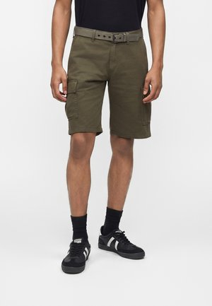 Άνδρας που φοράει ελαιδί cargo σορτς με ζώνη, μαύρες κάλτσες και μαύρα αθλητικά παπούτσια με λευκές ρίγες, στέκεται σε απλό λευκό φόντο.