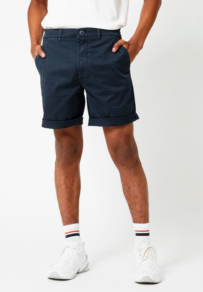 Shorts en coton bleu marine avec revers, dotés de poches avant et d'une fermeture à bouton sur le devant. Portés avec des baskets blanches et des chaussettes rayées.