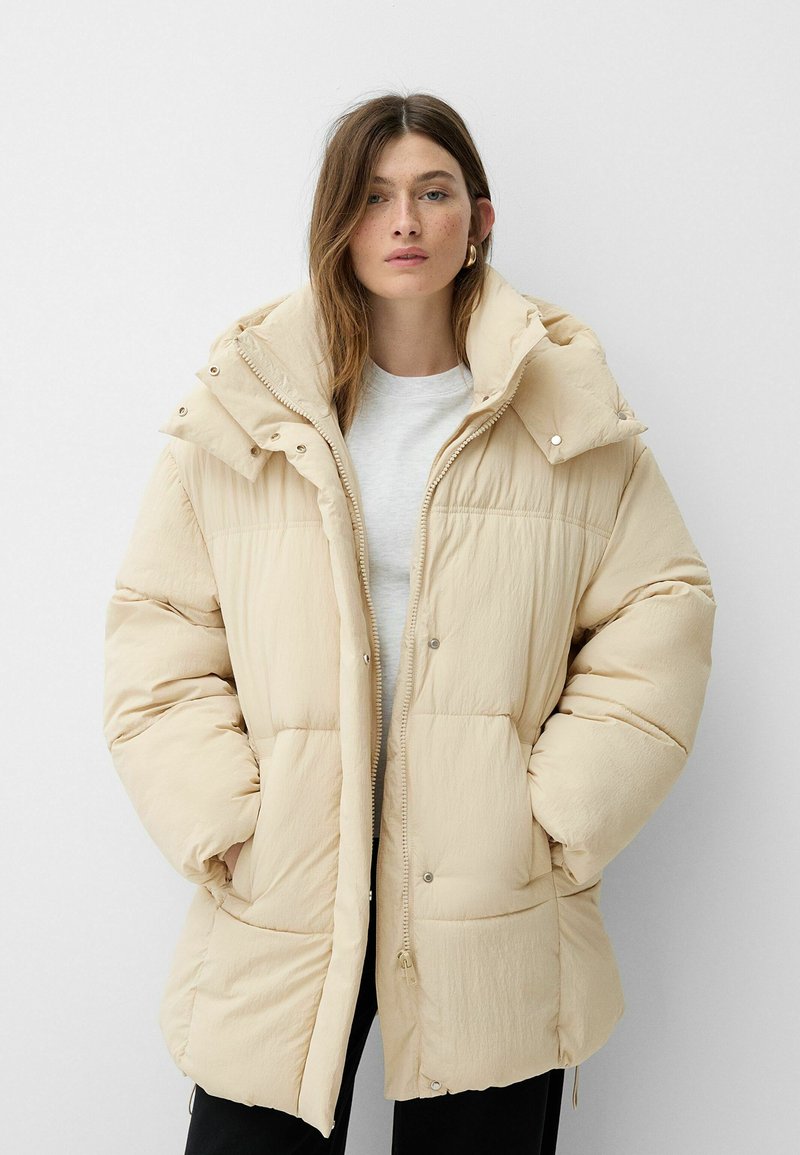 Giubbotto imbottito beige con colletto alto, chiusura a zip e bottoni a pressione. Presenta ampie tasche e un tessuto morbido e testurizzato.