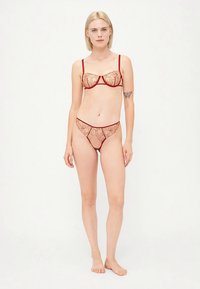 Ženska s kratkimi blond lasmi, ki stoji bosa v prosojnem bež spodnjem perilu z rdečim cvetličnim vezenjem in tankimi rdečimi naramnicami na bež ozadju.