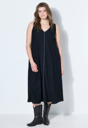 Junge Frau mit lockerem welligem Haar, die ein schwarzes ärmelloses Midikleid mit weißen Nähten und schwarzen Cowboy-Stiefeln trägt, steht vor einer weißen Wand.