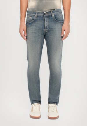 Faded blue denim jeans with a slim fit, five-pocket design, and subtle stitching details, worn with cream-colored sneakers.