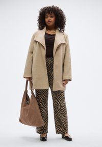 Cappotto oversize in lana beige, camicia nera, pantaloni a gamba larga a righe marroni e nere, borsa in camoscio marrone, scarpe basse nere. Materiali texture.