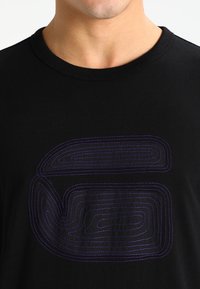 Mustan puuvillan t-paita, jossa on suuri, kaareva violetilla ommeltu logomuotoilu edessä ja klassinen pyörökaula-aukko.