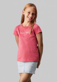 Camiseta de algodón rosa con mangas cortas y cuello redondo, que presenta el logo blanco de "Calvin Klein" en el pecho. Combinada con una falda mini de mezclilla.