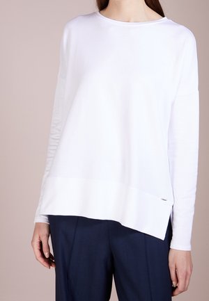 Žena v volném bílém topu s dlouhým rukávem a širokým lemem, v námořnických kalhotách na míru, stojící na neutrálním pozadí.