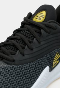Κοντινό πλάνο ενός μαύρου αθλητικού παπουτσιού με πλεκτό ύφασμα, μαύρα κορδόνια, κίτρινο λογότυπο στη γλώσσα και στο πλάι, και λευκή υφασμένη σόλα.
