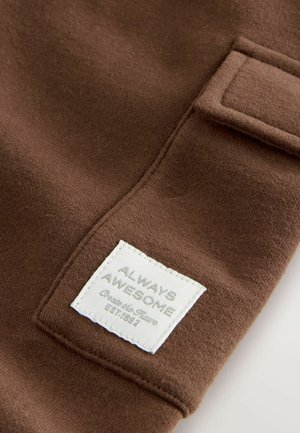 Barna anyagú, puha tapintású textil, amelyen egy négyzet alakú fehér címke található, rajta szürke betűkkel a "FOLYAMATOSAN CSODÁS" felirattal. Látható varrásokkal kiemelve.