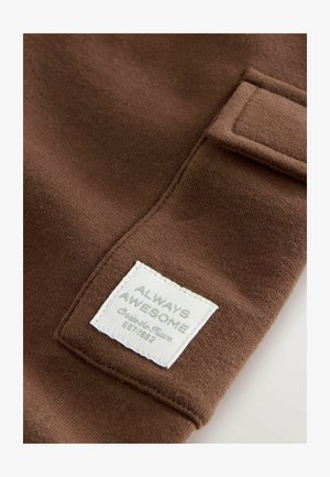 Barna anyagú, puha tapintású textil, amelyen egy négyzet alakú fehér címke található, rajta szürke betűkkel a "FOLYAMATOSAN CSODÁS" felirattal. Látható varrásokkal kiemelve.