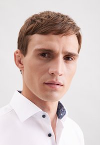 Witte button-up blouse met een patroon op de blauwe kraag en zwarte knopen, met een fitted design en een zachte textuur, met de nadruk op het detail van de kraag.