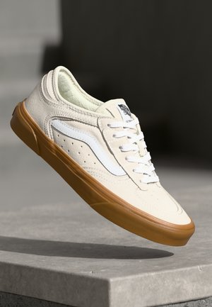 Beige suede sneaker with white accents, featuring a perforated design and a gum rubber sole. Laced with white shoelaces.