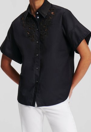 Chemise noire à manches courtes avec un col boutonné, présentant un design découpé complexe sur la poitrine et une texture de tissu lisse.