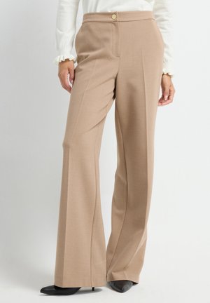Beige wijde broek met hoge taille en knoopdetails, gedragen met zwarte puntige hakken en een witte blouse met geribbelde manchetten.