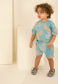 Ein Outfit für Kleinkinder bestehend aus einem kurzärmligen türkisfarbenen Hemd mit orangenen Blättern und passenden Shorts. Getragen mit grauen Sandalen.