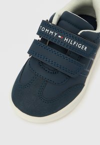 Tmavě modrá teniska s pásky na suchý zip a kontrastními bílými detaily. Obsahuje prošité ozdobné prvky a logo na pásku. Gumová podrážka.