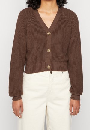Personne portant un cardigan en maille marron boutonné à manches longues et un pantalon crème taille haute, les mains détendues sur les côtés.