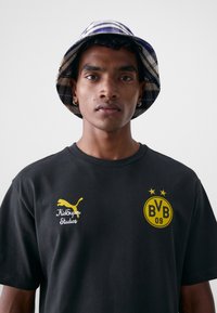 Svart bomull t-skjorte med gult Puma-logo og "KidSuper Studios" tekst. Laget rutete bøttehatt i blå og beige toner.