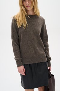 Barna gyapjú pulóver hosszú ujjakkal és bordázott szegéllyel, fekete bőr szoknya fölött viselve. A modell egy sötétbarna kézitáskát tart.