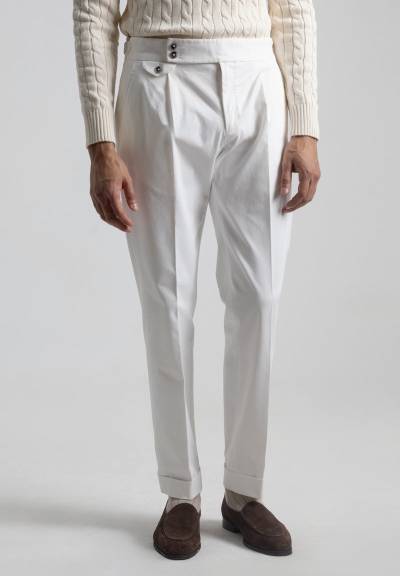 Pantalones de algodón blancos con un frontal plisado, que cuentan con dos botones negros en la cintura y puños. Unos zapatos de gamuza marrón completan el look.