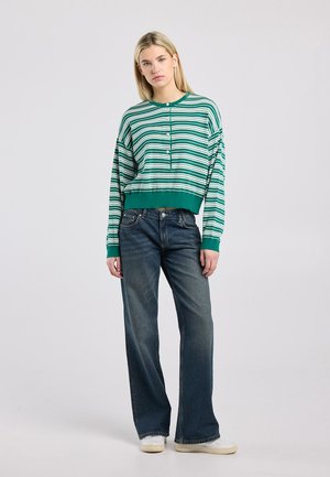 Jonge vrouw staand, gekleed in een groen-wit gestreept shirt met lange mouwen, donkere jeans met wijde pijpen en witte sneakers, neutrale achtergrond.