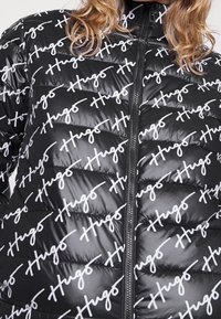 Sort pufjakke med hvide "Hugo" logoer, quiltet design, høj krave og lynlås foran. Glat, blank tekstur.