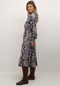 Suknja z dolgimi rokavi v mornarsko modri barvi z bež vzorcem paisley, elastičnim pasom in večplastnim krilom. Ujemite jo z rjavimi čevlji do kolen.