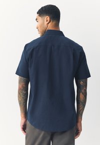 Camicia blu navy a maniche corte con bottoni, tessuto liscio, colletto standard e orlo curvo, vista posteriore.