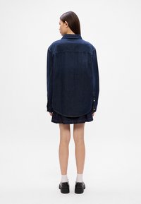 Τζιν πουκάμισο σε σκούρο μπλε χρώμα, με μυτερό γιακά, μπροστινό κλείσιμο με κουμπιά και κυρτή βάτα. Ο ιστός παρουσιάζει λεία υφή με ελάχιστες ραφές.