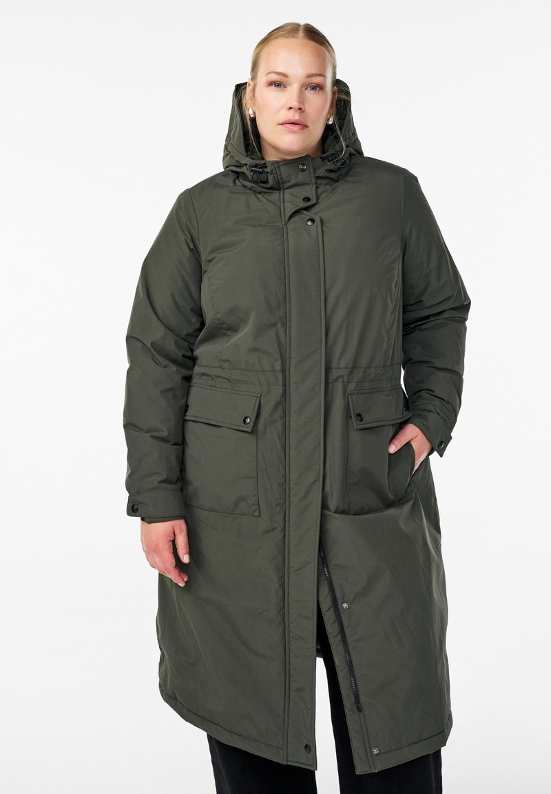 Ženska v dolgem, olivno zelenem zimski plašču s kapuco, sprednjimi žepi s poklopcem in gumbi, ki stoji pred belo ozadjem.