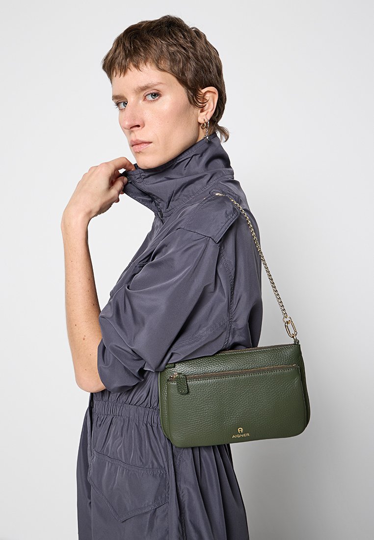 Grüne Lederhandtasche mit strukturiertem Finish, rechteckiger Form, vorderer Reißverschlusstasche und silberner Kettenriemen. Logo auf der Vorderseite sichtbar.